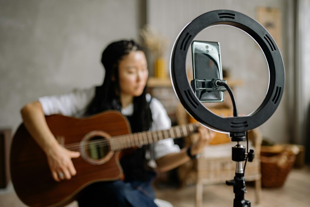 Focused woman playing guitar during a live stream with a smartphone in a ring light indoors.