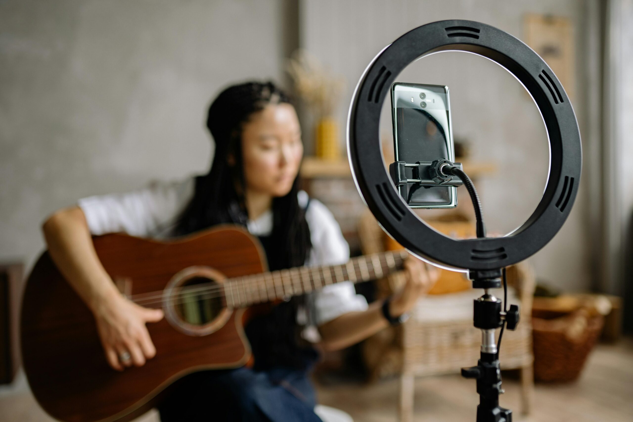Focused woman playing guitar during a live stream with a smartphone in a ring light indoors.
